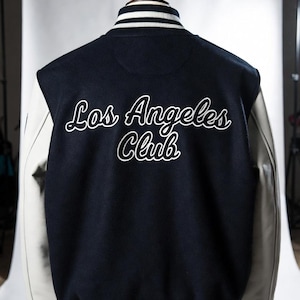 May include: Navy blue and white varsity jacket with the text "Los Angeles Club" in white script on the back. The jacket has white leather sleeves and striped cuffs and collar. The jacket is displayed on a mannequin.