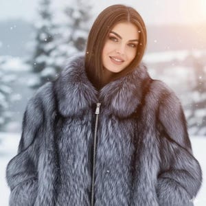 Puede incluir: Un abrigo de piel plateado y gris con cierre de cremallera. El abrigo tiene una textura suave y un cuello ancho. El fondo muestra una escena nevada al aire libre.