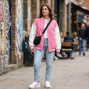 Op de afbeelding: Een persoon draagt een roze en witte varsity-jas, lichtblauwe jeans en witte sneakers. Ze draagt een zwarte crossbodytas. De achtergrond omvat een bakstenen muur met graffiti en een straatbeeld.