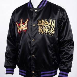 May include: Black satin bomber jacket with gold and red embroidered crown and "URBAN KINGS" text. The jacket has purple and black striped trim on the collar, cuffs, and hem. Gold snap closures complete the look.