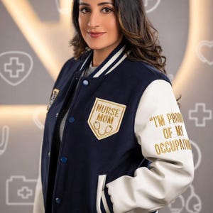 Może przedstawiać: Granatowo-biała kurtka typu letterman z napisem "NURSE MOM" i emblematem stetoskopu na piersi. Prawy rękaw ma napis "I'M PROUD OF MY OCCUPATION" złotymi literami. Kurtka ma białe rękawy i niebieskie guziki.
