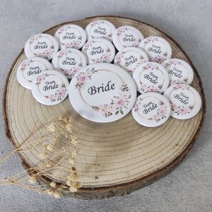 Könnte beinhalten: Ein Set mit 12 runden weißen Anstecknadeln mit Blumendesigns und dem Text "Team Bride". Eine größere Anstecknadel zeigt ein Blumendesign und den Text "Bride".