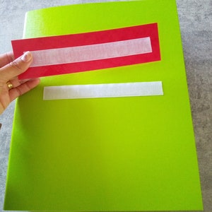 May include: A bright green notebook with a red rectangle and white velcro strip. A hand holds a red rectangle with a white velcro strip, positioned above the notebook. Another white velcro strip is attached to the notebook.