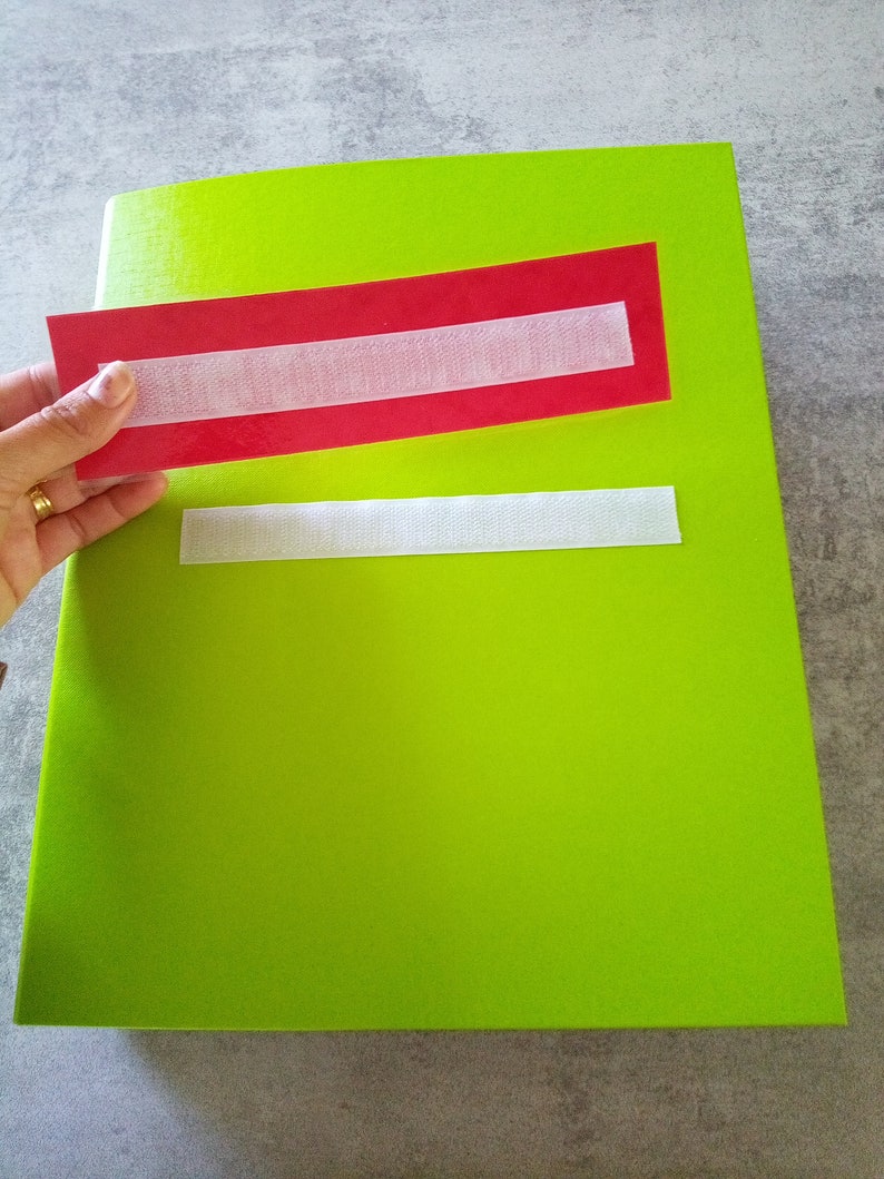 May include: A bright green notebook with a red rectangle and white velcro strip. A hand holds a red rectangle with a white velcro strip, positioned above the notebook. Another white velcro strip is attached to the notebook.