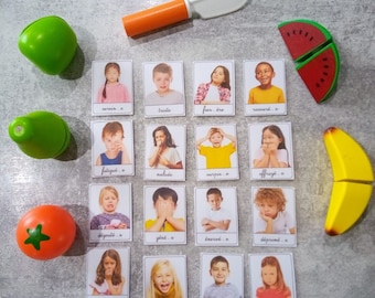 Puede incluir: Un conjunto de 16 tarjetas de emociones que muestran niños con diferentes expresiones faciales. Las tarjetas están etiquetadas con la emoción correspondiente en español. El conjunto también incluye una variedad de frutas y verduras de juguete, incluyendo un tomate rojo, una pera verde, una manzana verde, un plátano amarillo y una sandía roja.