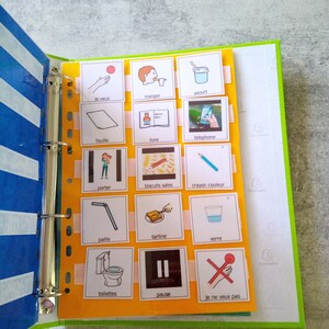 May include: A binder with a blue and white striped cover, holding pages with illustrated cards. The cards feature simple drawings and French words like "manger" (eat), "yaourt" (yogurt), and "pause". The binder is open, displaying the cards.