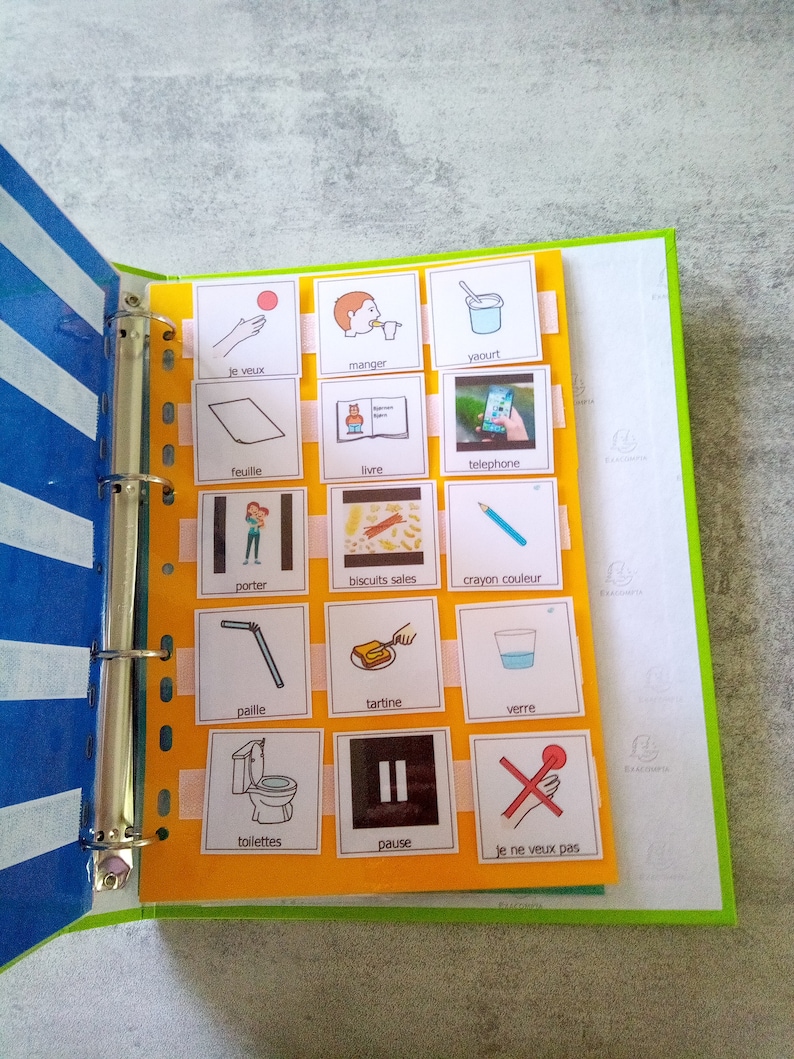 May include: A binder with a blue and white striped cover, holding pages with illustrated cards. The cards feature simple drawings and French words like "manger" (eat), "yaourt" (yogurt), and "pause". The binder is open, displaying the cards.