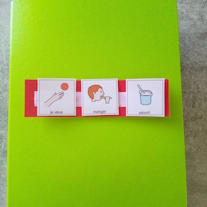 May include: A bright green notebook with a red and white strip across the front. The strip has three small images with text: a hand throwing a ball, a person eating, and a cup of yogurt. The text includes the words "je veux", "manger", and "yaourt".