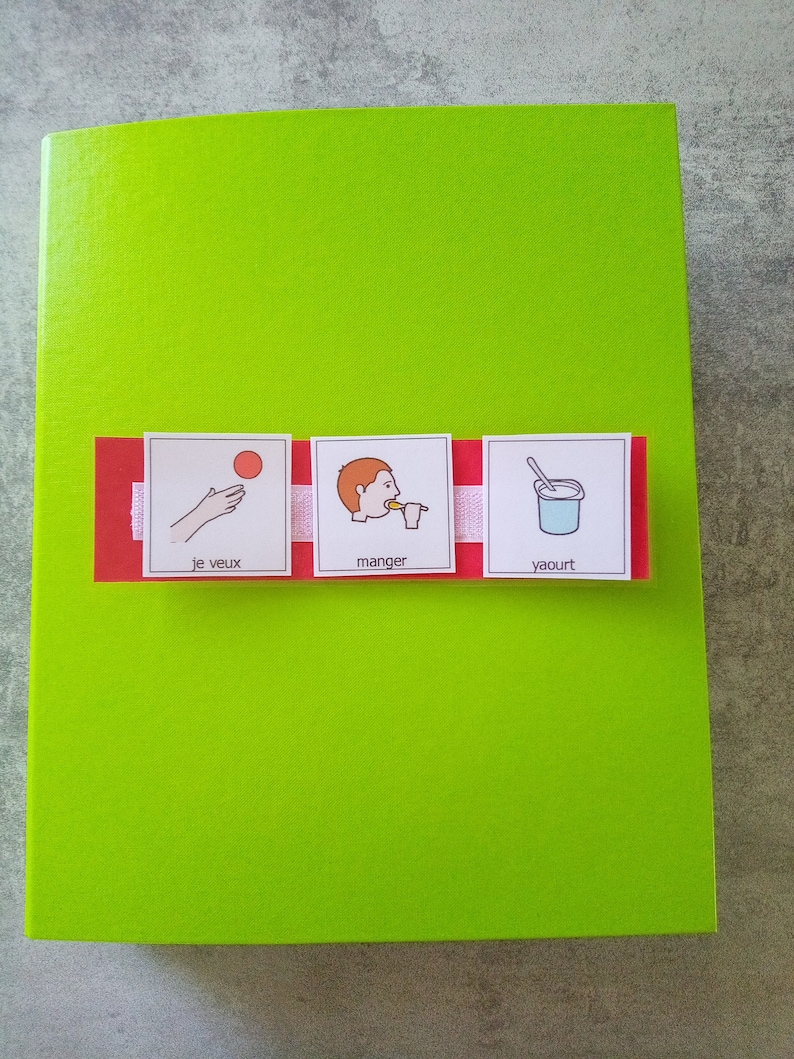 May include: A bright green notebook with a red and white strip across the front. The strip has three small images with text: a hand throwing a ball, a person eating, and a cup of yogurt. The text includes the words "je veux", "manger", and "yaourt".