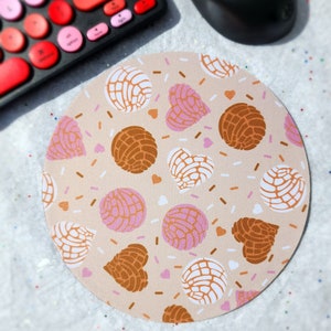 May include: A round mouse pad with a light brown background and a pattern of pink and brown concha pastries with white sprinkles and hearts.