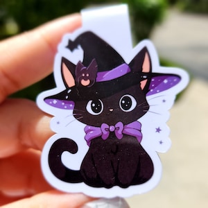 May include: A black cat wearing a purple witch hat with a small bat on top. The cat has a purple bow tie and is surrounded by small white stars.
