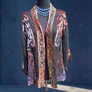 May include: Black open-front jacket with velvet floral patterns in red, purple, and pink. The jacket features gold embroidered trim along the edges and a blue beaded necklace. The jacket is displayed on a black mannequin.