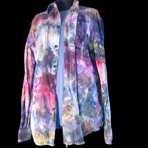 Może przedstawiać: Koszula z długim rękawem zapinana na guziki, ozdobiona żywym wzorem tie-dye w odcieniach r&oacute;żu, fioletu, błękitu i ż&oacute;łci. Koszula posiada klasyczny kołnierzyk, przednią listwę z ciemnymi guzikami oraz długie rękawy z mankietami, założona na gładką niebieską koszulkę.
