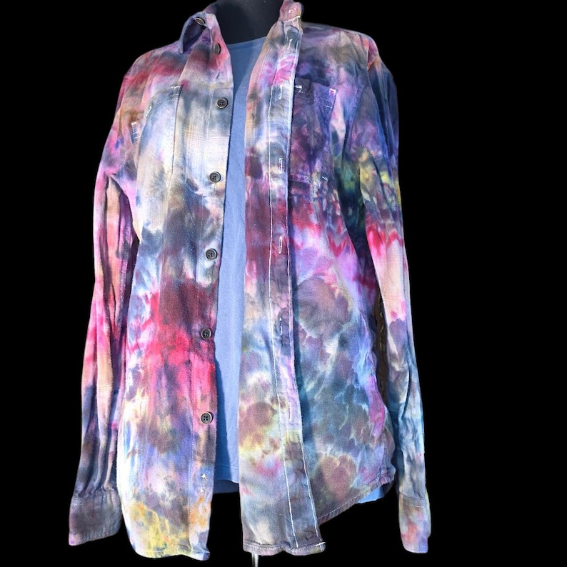 Może przedstawiać: Koszula z długim rękawem zapinana na guziki, ozdobiona żywym wzorem tie-dye w odcieniach r&oacute;żu, fioletu, błękitu i ż&oacute;łci. Koszula posiada klasyczny kołnierzyk, przednią listwę z ciemnymi guzikami oraz długie rękawy z mankietami, założona na gładką niebieską koszulkę.