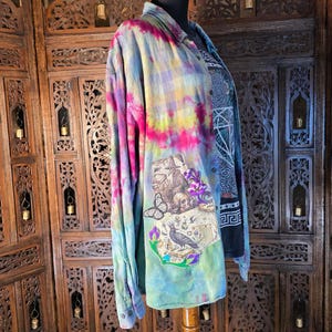 Könnte beinhalten: Ein langärmeliges Hemd mit Knopfleiste und einem lebendigen Batikmuster in Pink, Gelb und Blau. Auf der Vorderseite befindet sich ein rechteckiger Aufnäher mit detaillierten Illustrationen eines Raben, eines Schmetterlings, eines Schädels und eines Zauberers.
