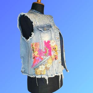 May include: A light blue denim vest featuring a frayed hem and armholes, embellished with a vibrant floral patch in shades of pink, yellow, and green on the front, along with silver-toned metal studs along the collar and shoulder area.