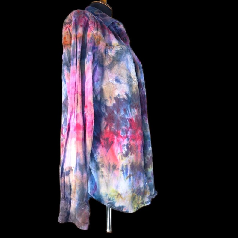 Może przedstawiać: Koszula z długim rękawem zapinana na guziki, ozdobiona żywym wzorem tie-dye z mieszanką odcieni r&oacute;żu, fioletu, błękitu i ż&oacute;łtymi akcentami. Koszula posiada kołnierzyk, pojedynczą kieszeń na lewej piersi oraz mankiety zapinane na guziki.