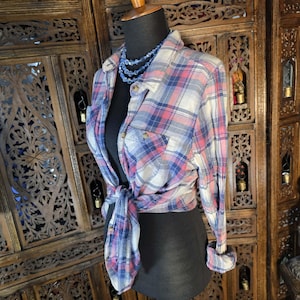 May include: A plaid button-down shirt in shades of red, blue, and white is tied at the waist on a mannequin. The long-sleeved shirt has a classic collar and two chest pockets. A blue beaded necklace is worn with the shirt.