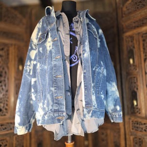 May include: A blue denim jacket with a bleached tie-dye pattern featuring a fold-over collar, long sleeves, and a button-front closure. Underneath, a grey button-down shirt with a dark star print and a black graphic t-shirt are layered.