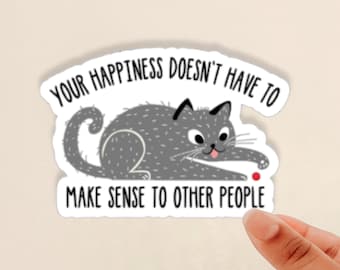 Puede incluir: Un gato gris con la lengua rosa está jugando con una pelota roja. El texto en la pegatina dice "Tu felicidad no tiene que tener sentido para los demás."