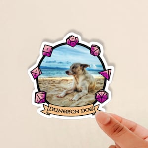 May include: A sticker featuring a cartoon dog with pink dice around it. The dog is lying on a beach with the text "DUNGEON DOG" below it.