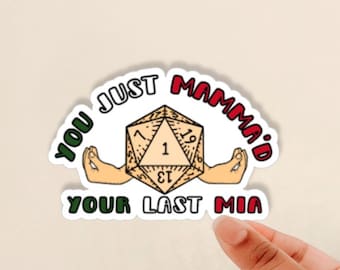 May include: A white sticker with a red, green, and white border. The sticker features a cartoon image of a 20-sided die with arms outstretched. The text on the sticker reads "You Just Mamma'd Your Last Mia".