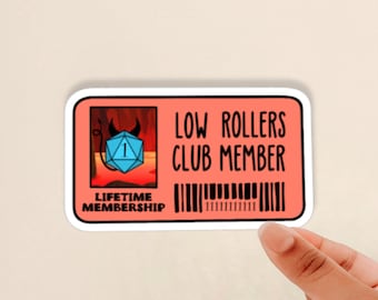 May include: A red and white sticker with the text "Low Rollers Club Member" and a cartoon image of a blue D20 die with a 1 showing. The die is in a red and black background with a devil's tail. The text "Lifetime Membership" is below the die.