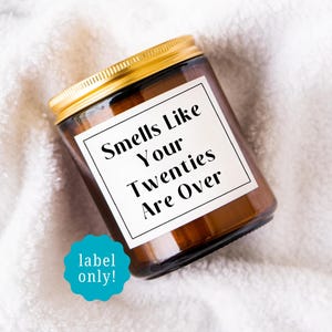May include: A brown glass candle jar with a gold lid. The label reads "Smells Like Your Twenties Are Over" in black text. A teal sticker at the bottom says "label only!". The candle sits on a white, textured surface.