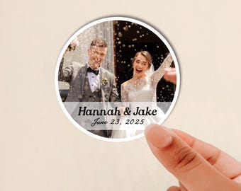 May include: A circular sticker featuring a photograph of a couple in wedding attire with confetti falling around them. The text "Hannah & Jake" and "June 23, 2025" is printed in black across a semi-transparent white horizontal band.
