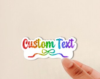 May include: A die-cut sticker featuring the words Custom Text in a rainbow glitter gradient. Below the text is a decorative ribbon bow graphic, also rendered in a matching rainbow glitter finish, with a white border surrounding the entire design.