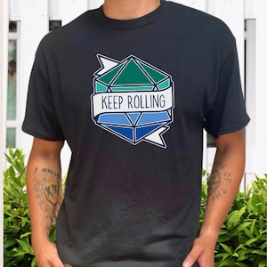 Peut inclure: T-shirt noir avec un motif géométrique bleu et vert, et les mots "KEEP ROLLING" sur une bannière. Le motif ressemble à un dé à multiples facettes. Un vêtement décontracté.