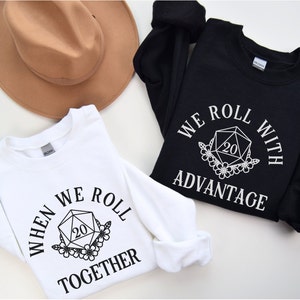 Op de afbeelding: Twee witte en zwarte sweatshirts met een afbeelding van een twintigzijdige dobbelsteen met bloemen en de tekst "When we roll together" en "We roll with advantage".