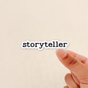 May include: A white sticker with the word "storyteller" in a black, sans-serif font. The sticker is held by a person's hand against a light beige background. The sticker has a white border.