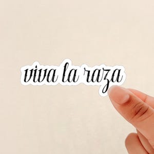May include: A white sticker with the phrase "viva la raza" in black cursive script. The sticker is being held by a hand against a light beige background. The text is outlined in black.