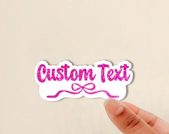 May include: A die-cut sticker featuring the words Custom Text in a bright pink glitter texture. The design includes a decorative pink glitter ribbon flourish underneath the text, all set against a clean white background border.