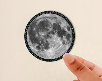 May include: A circular sticker featuring a detailed, high-contrast photographic image of the full moon in shades of gray, white, and black. The moon is surrounded by a thin, dark border filled with small, multicolored, reflective glitter particles.
