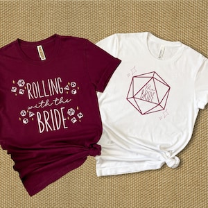 May include: Two t-shirts, one maroon and one white, with a dice theme. The maroon shirt says "Rolling with the Bride" and has small dice graphics. The white shirt has a large dice graphic with "The Bride" inside.