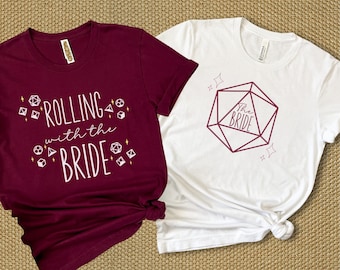 May include: Two t-shirts, one maroon and one white, with a dice theme. The maroon shirt says "Rolling with the Bride" and has small dice graphics. The white shirt has a large dice graphic with "The Bride" inside.