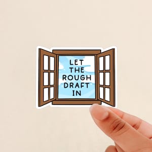 May include: A sticker featuring a brown window frame with open window panes. The window reveals a blue sky with clouds and the text "LET THE ROUGH DRAFT IN". The sticker is held by a person's hand.