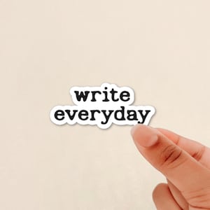 May include: A white sticker with the words "write everyday" in a black, bold font. The sticker is held by a person's fingers against a light beige background. The sticker has a white border.