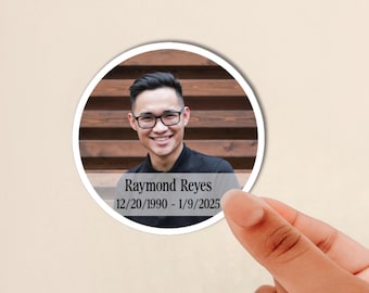 May include: A circular sticker featuring a photograph of a smiling person wearing glasses and a black shirt. The sticker includes a semi-transparent gray overlay at the bottom with the text Raymond Reyes and the dates 12/20/1990 - 1/9/2025.