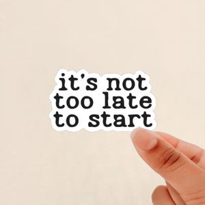 May include: A white sticker with the phrase "it's not too late to start" in black, bold, sans-serif font. The sticker is held by a hand against a light beige background.