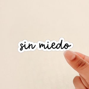 May include: A white sticker with the phrase "sin miedo" in black cursive script. The sticker is held by a hand against a light beige background. The phrase translates to "without fear".