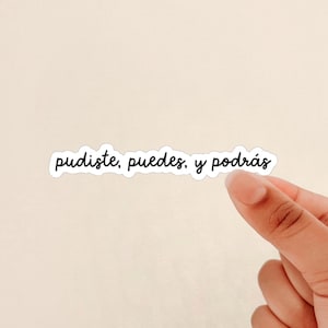 May include: A white sticker with the words "pudiste, puedes, y podrás" in black cursive font. The sticker is held by a hand against a light beige background.