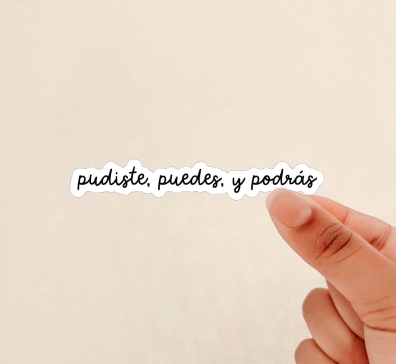 May include: A white sticker with the words "pudiste, puedes, y podr&aacute;s" in black cursive font. The sticker is held by a hand against a light beige background.