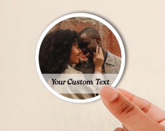 May include: A circular sticker featuring a photograph of two people in an intimate embrace against a textured reddish background. The sticker has a white border and a semi-transparent horizontal band across the bottom with the text Your Custom Text.