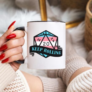 May include: White ceramic mug with a black handle and rim. The mug features a geometric design in the colours of the transgender flag, with the text "KEEP ROLLING" on a banner. The design includes the numbers 2, 8, 14, 20, and 9.