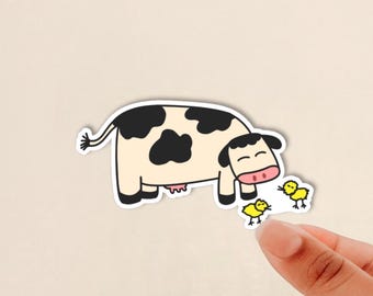 May include: A die-cut sticker featuring a cartoon cow with a cream-colored body and black spots. The cow has a pink snout and is positioned next to two small, bright yellow chicks. The design has a thick white border around the entire illustration.