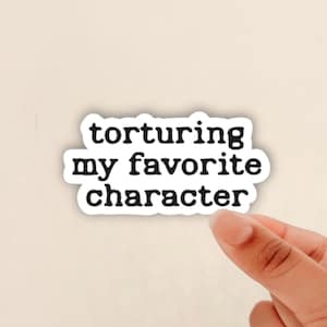 May include: A white sticker with the text "torturing my favorite character" in a black, bold font. The sticker is held by a person's fingers against a neutral background.