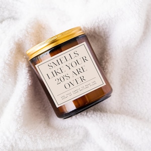 May include: A brown glass candle with a gold lid and a white label that reads "Smells Like Your 20's Are Over" and "Use this candle to mask the scent of your rotting flesh."